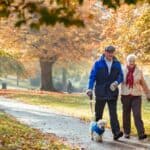 Senior couple are walking their dog through a public park in Autumn.